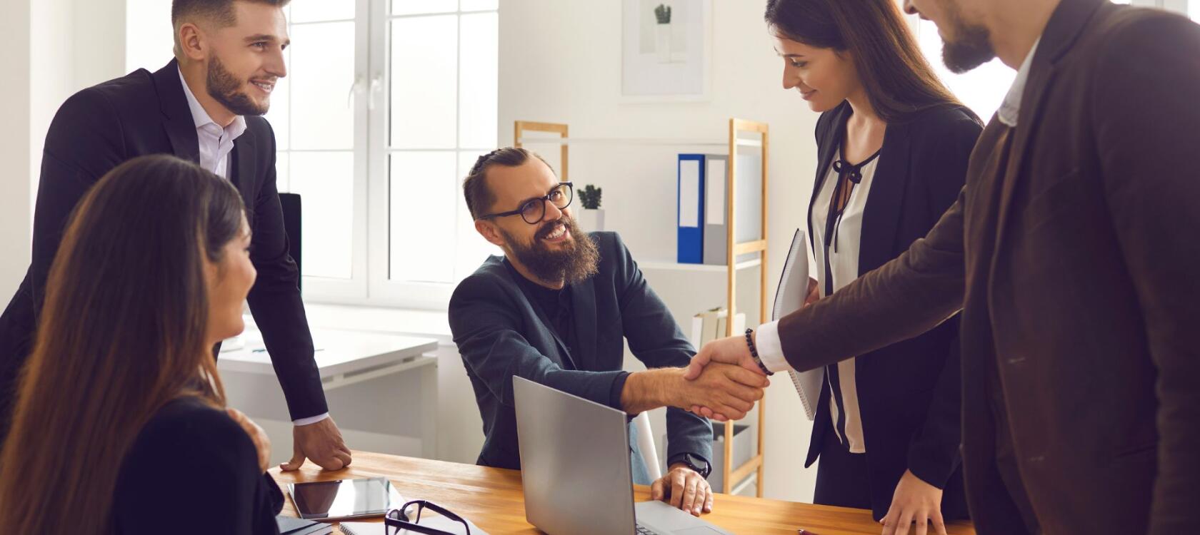 A group of happy people in an office setting, setting up a deal with a partner, one of the people is shaking hands with the guest, showing partnership.