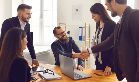 A group of happy people in an office setting, setting up a deal with a partner, one of the people is shaking hands with the guest, showing partnership.