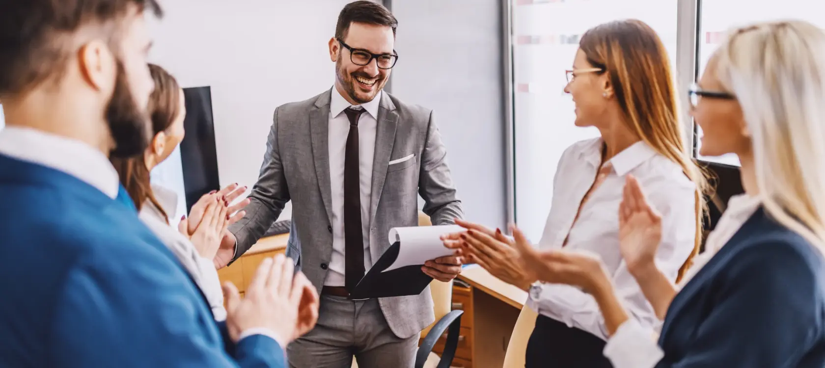 A photo of an office cheering their team member after a successful job done or a goal achieved. The colleagues are clapping while the male colleague wearing a gray suite and glasses is happily announcing success by reading the charts. 