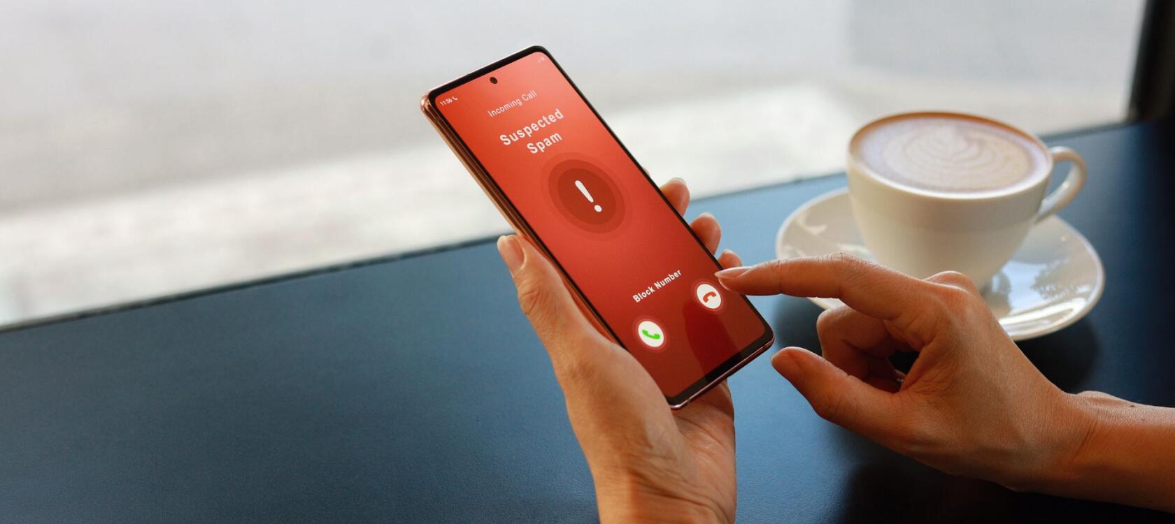 Female hands using a cell phone in a cafe setting. The phone display shows "Suspected Scam" on a red screen and the user's finger is reluctant whether to pick up the call or not. This is a cover image for Heksagon's Telecom Fraud Guide article. 