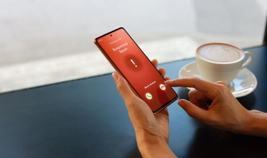 Female hands using a cell phone in a cafe setting. The phone display shows "Suspected Scam" on a red screen and the user's finger is reluctant whether to pick up the call or not. This is a cover image for Heksagon's Telecom Fraud Guide article. 
